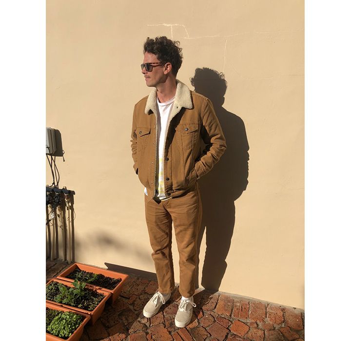 Photo of David Davey standing against a cream wall outside. He's wearing a brown corduroy jacket, a white tee, and tan 514s with tan sneakers.
