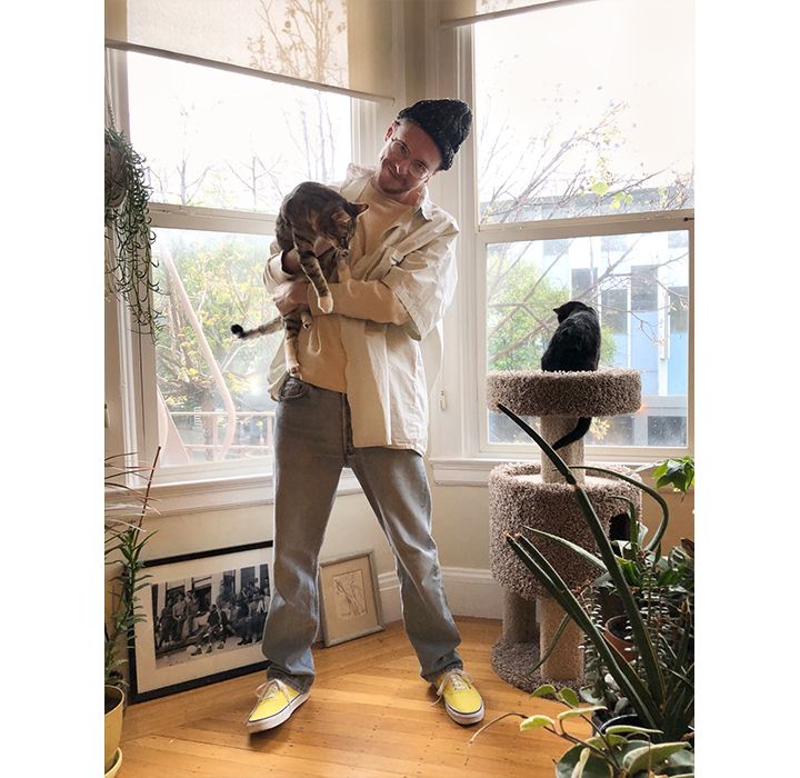 Photo of Ezra Price in his apartment. He's holding his cat and smiling.