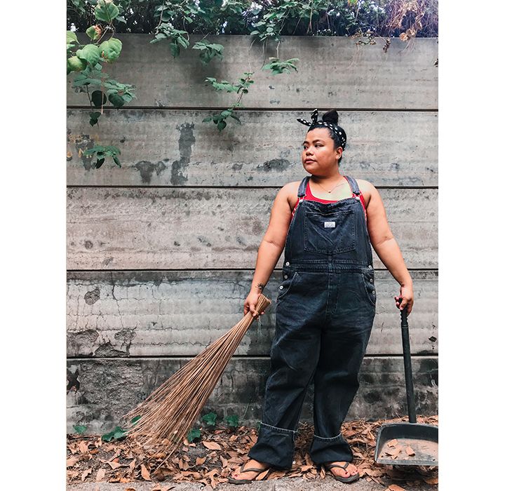 Photo of Jhae Lasaca standing outside in front of a fence. She's wearing dark Levi's overalls over a red tank top with flip flops.