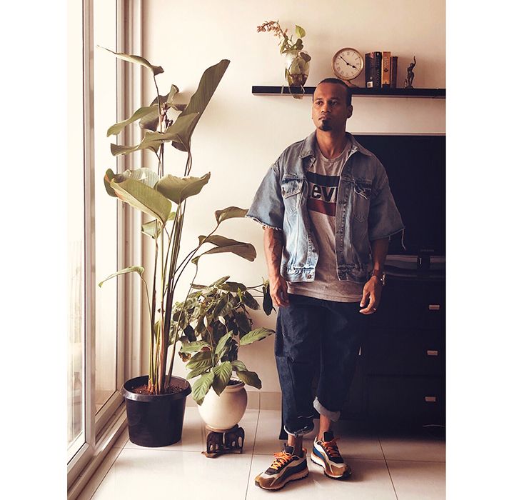 Photo of Ramu Mohan standing by a window inside his home. He is wearing a Levi's logo tee, a Levi's Trucker Jacket with cut-off, frayed sleeves, cuffed jeans, and chunky, colorful sneakers.