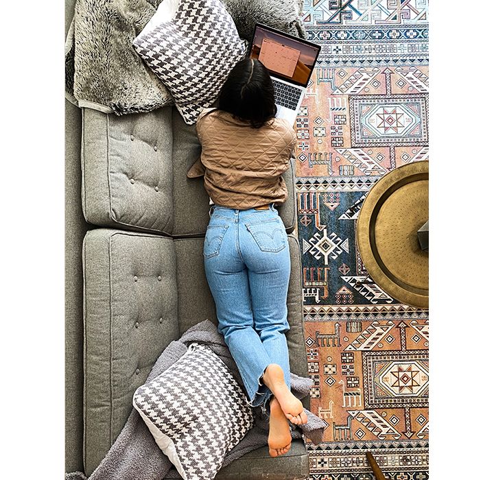 Aerial shot of Zoe Chrysochoos lying on her couch on her stomach with her laptop open. She is wearing a brown top and Levi's jeans.