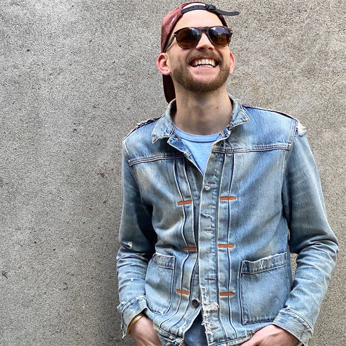 Photo of Austin McCune standing against a concrete wall. He is wearing a baseball hat backwards, sunglasses, and a faded Levi's Trucker Jacket.