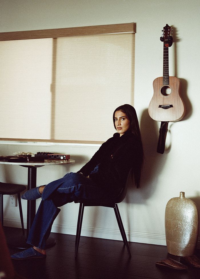 Portrait of ABIR sitting in a chair with a guitar hanging on the wall behind her.