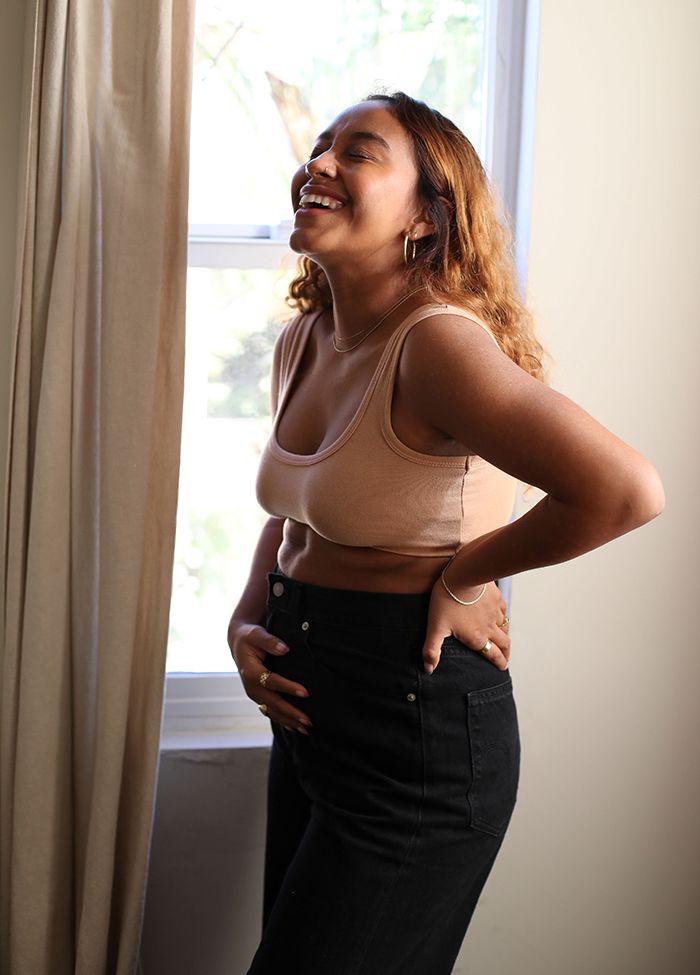 Photo of Evelynn Escobar-Thomas holding her pregnant belly and throwing her head back in laughter. She is wearing a peach sports bra and black jeans.